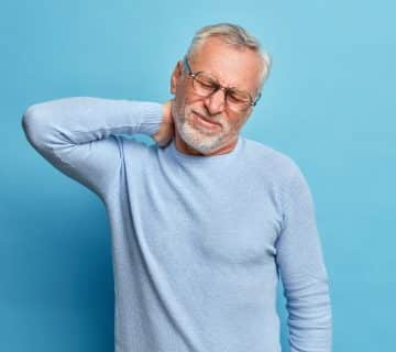 Aged exhausted bearded european man touches neck suffers from pain neck tilts head grimaces from painful feelings needs massage dressed long sleeved jumper isolated blue wall 360x320